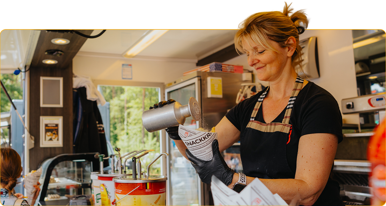 Medewerkster in een snackwagen strooit smaakmaker over een portie verse frietjes terwijl ze de bestelling bereidt.